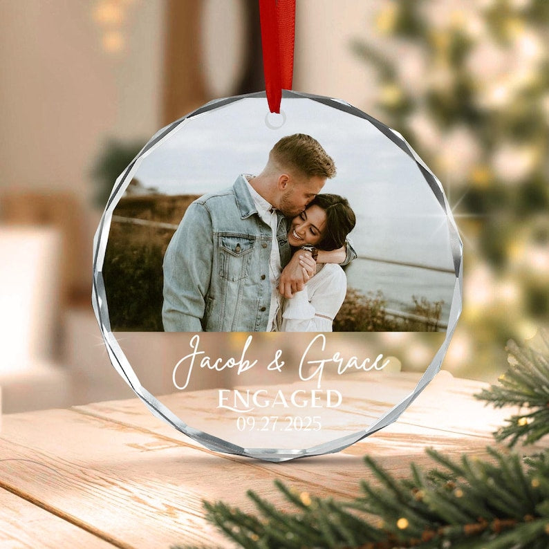 A circular glass ornament featuring a photograph of a couple embracing outdoors. The text "Jacob & Grace" is printed in white script, followed by "ENGAGED" and the date "09.27.2025" in white lettering.