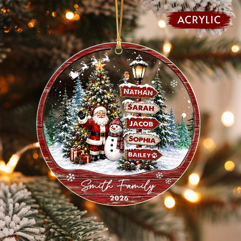 A round acrylic Christmas ornament featuring a winter scene with Santa Claus, a snowman, and a decorated tree. A street sign post displays the names Nathan, Sarah, Jacob, Sophia, and Bailey.