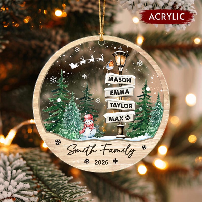 A round acrylic Christmas ornament featuring a winter scene with green pine trees, a snowman, a street lamp with signs reading MASON, EMMA, TAYLOR, and MAX with a paw print, a flying sleigh, and the text Smith Family 2026.