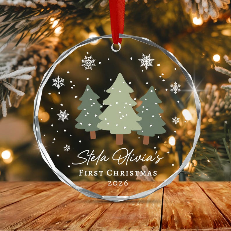 A circular clear acrylic ornament with faceted edges, featuring a printed design of three green pine trees and white snowflakes. The text reads Stela Olivia's FIRST CHRISTMAS 2026. A red ribbon is attached to the top for hanging.