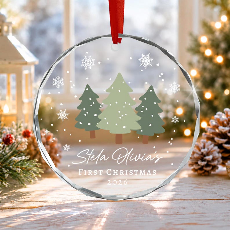 A circular clear glass ornament with a red ribbon hanger, featuring a printed design of three green pine trees, white snowflakes.