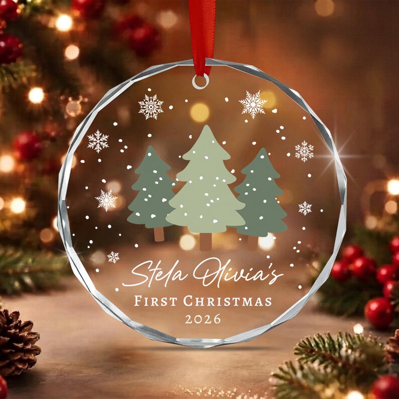 A circular clear glass ornament with a faceted edge, featuring a printed design of three green pine trees and white snowflakes. The text reads Stela Olivia's FIRST CHRISTMAS 2026. A red ribbon is attached to the top for hanging.