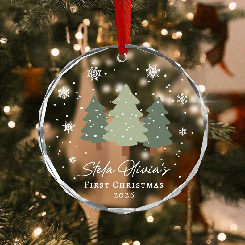 A circular clear glass Christmas ornament featuring a printed design of three green pine trees and white snowflakes. The text reads Stela Olivia's FIRST CHRISTMAS 2026 in white script and block lettering.