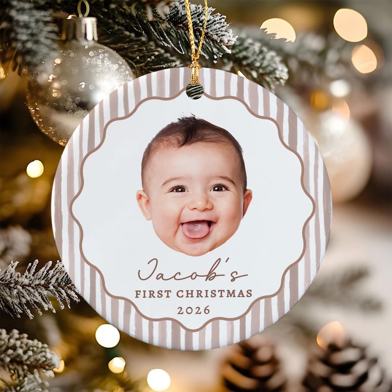 A round ceramic Christmas ornament featuring a photograph of a smiling baby in the center. The ornament has a white background with light brown vertical stripes and a scalloped border. Text reads Jacob's FIRST CHRISTMAS 2026.