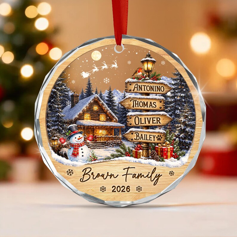 A round, faceted glass Christmas ornament featuring a winter scene with a log cabin, a snowman, and a signpost with the names Antonino, Thomas, Oliver, and Bailey.