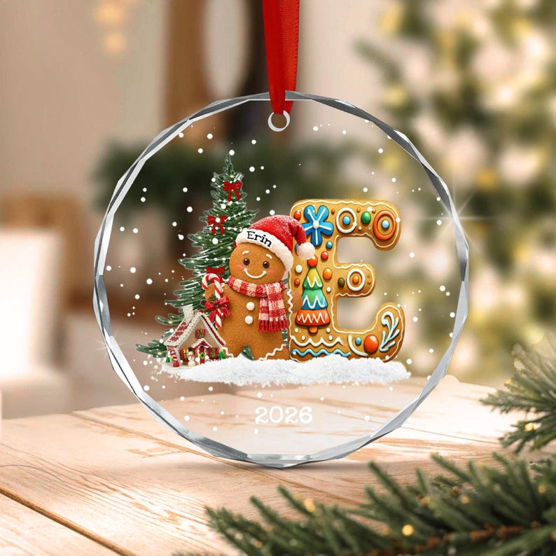 A circular clear glass Christmas ornament featuring a gingerbread person wearing a red hat with the name Erin, a green Christmas tree, a small house, and a large letter E decorated with icing.
