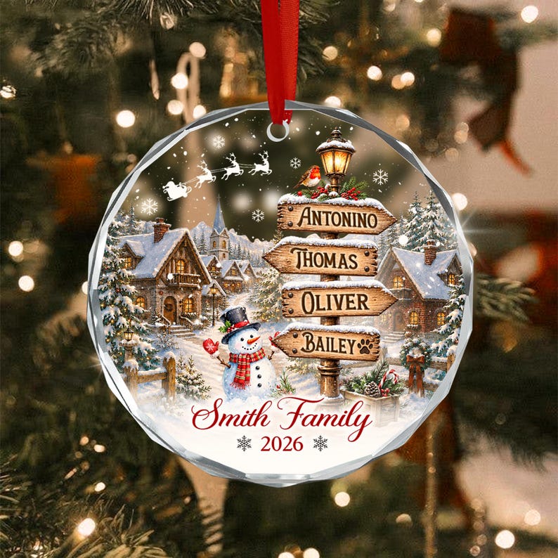 A round, faceted glass Christmas ornament featuring a winter village scene with a snowman, a signpost with the names Antonino, Thomas, Oliver, and Bailey, and the text Smith Family 2026, suspended by a red ribbon.