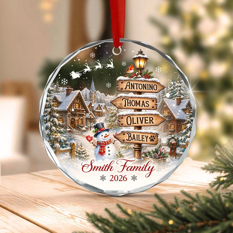 A round, faceted glass Christmas ornament featuring a winter village scene with a snowman, a street sign post with the names Antonino, Thomas, Oliver, and Bailey, and the text Smith Family 2026, suspended by a red ribbon.