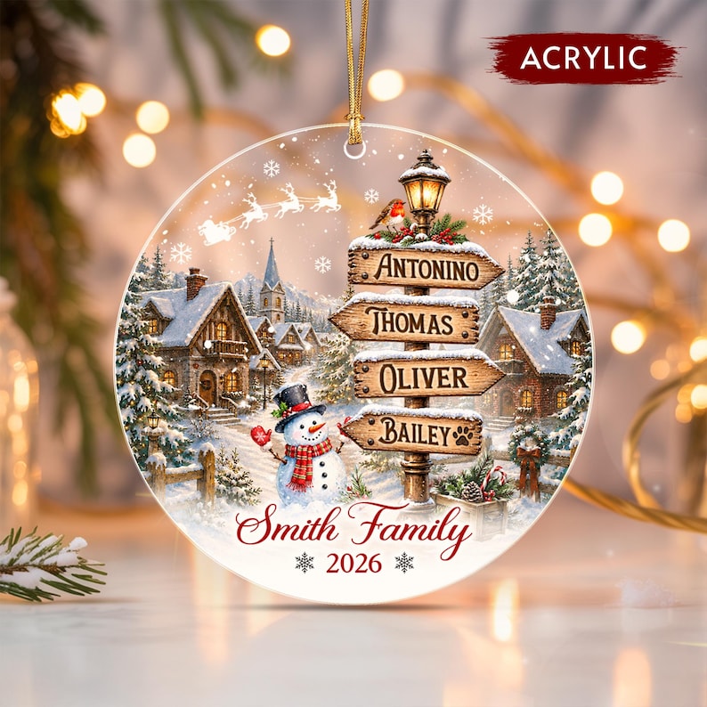 A round acrylic Christmas ornament featuring a snowy village scene with a snowman, a signpost with the names Antonino, Thomas, Oliver, and Bailey, and the text Smith Family 2026. A gold string is attached to the top for hanging.