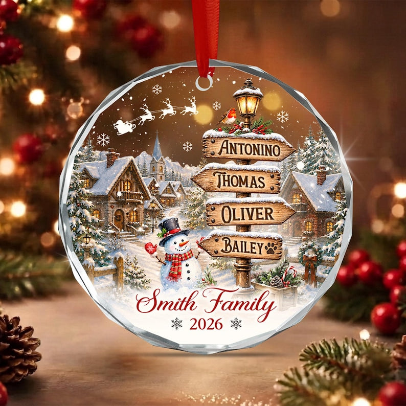 A round, faceted glass Christmas ornament featuring a winter village scene with a snowman, a signpost with the names Antonino, Thomas, Oliver, and Bailey, and the text Smith Family 2026, suspended by a red ribbon.