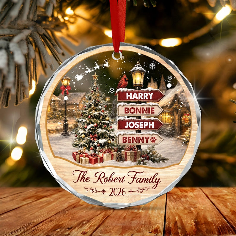 A round, faceted glass Christmas ornament hanging by a red ribbon. The design features a snowy village scene with a decorated tree, a street lamp, and a signpost with the names Harry, Bonnie, Joseph, and Benny.