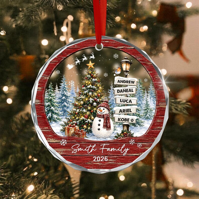 A round Christmas ornament with a red wooden-textured border featuring a snowy forest scene, a decorated Christmas tree, a snowman with a red scarf, and a street sign listing the names Andrew, Daneal, Luca, Ariel, and Kobe.