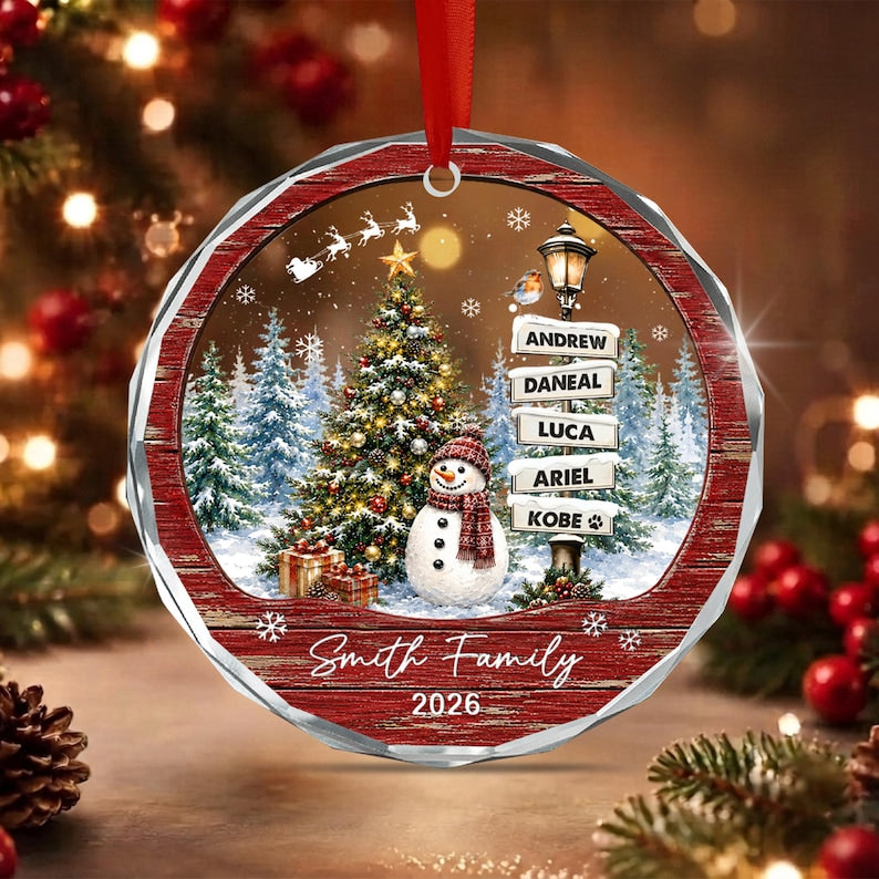 A round, red-framed Christmas ornament featuring a winter scene with a decorated tree, a snowman wearing a red scarf, and a street sign listing the names Andrew, Daneal, Luca, Ariel, and Kobe.
