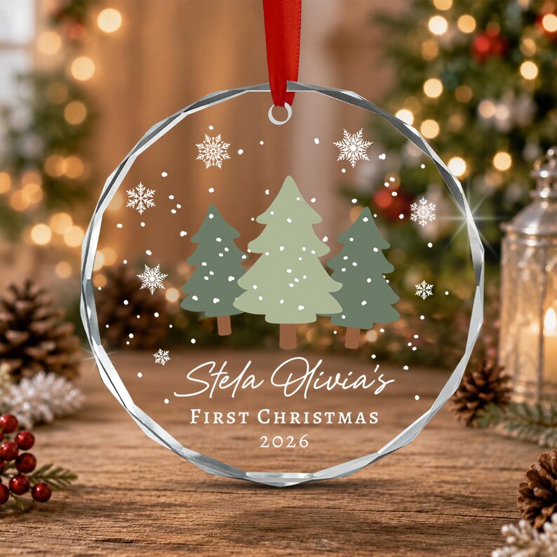 A circular, faceted glass Christmas ornament hanging from a red ribbon. The design features three stylized green pine trees, white snowflakes, and the printed text: Stela Olivia's FIRST CHRISTMAS 2026.