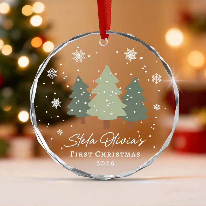 A circular clear glass ornament with faceted edges, featuring a printed design of three green pine trees, white snowflakes, and the text Stela Olivia's FIRST CHRISTMAS 2026. A red ribbon is attached to the top for hanging.