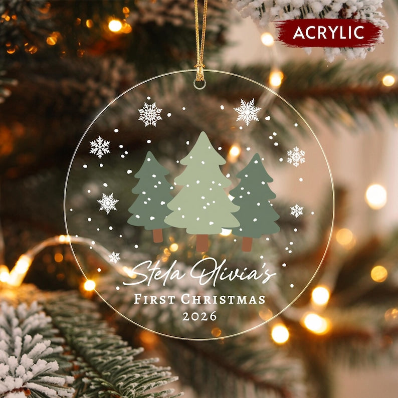 A round clear acrylic Christmas ornament featuring three illustrated pine trees in shades of green and brown, surrounded by white snowflakes. The text reads Stela Olivia's FIRST CHRISTMAS 2026 in white script and block lettering.
