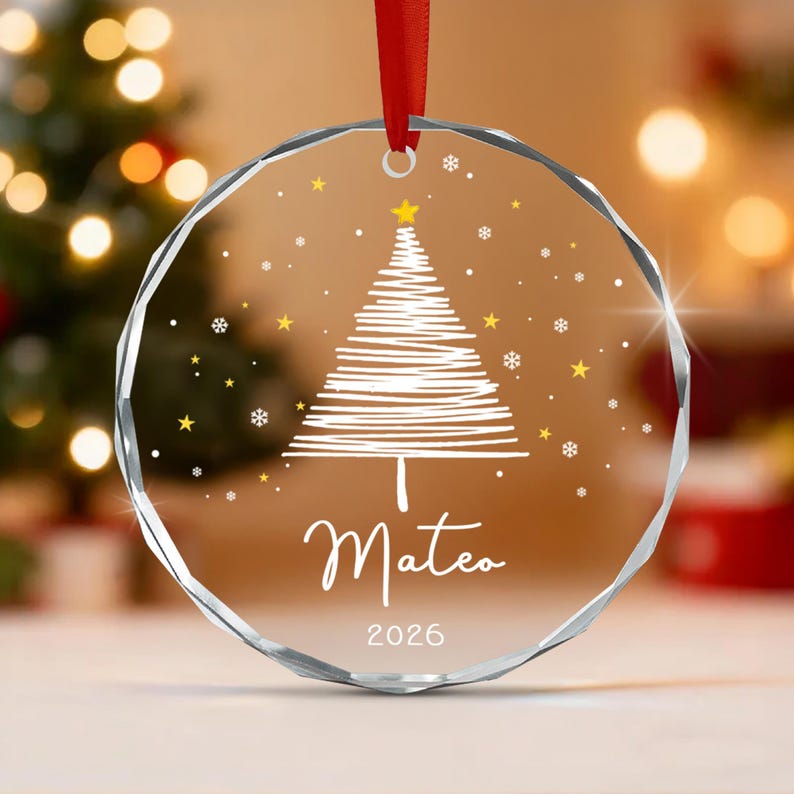 A circular clear glass ornament with faceted edges, featuring a white etched sketch of a Christmas tree, gold stars, and white snowflakes. The name Mateo is printed in white script above the year 2026, suspended by a red ribbon.
