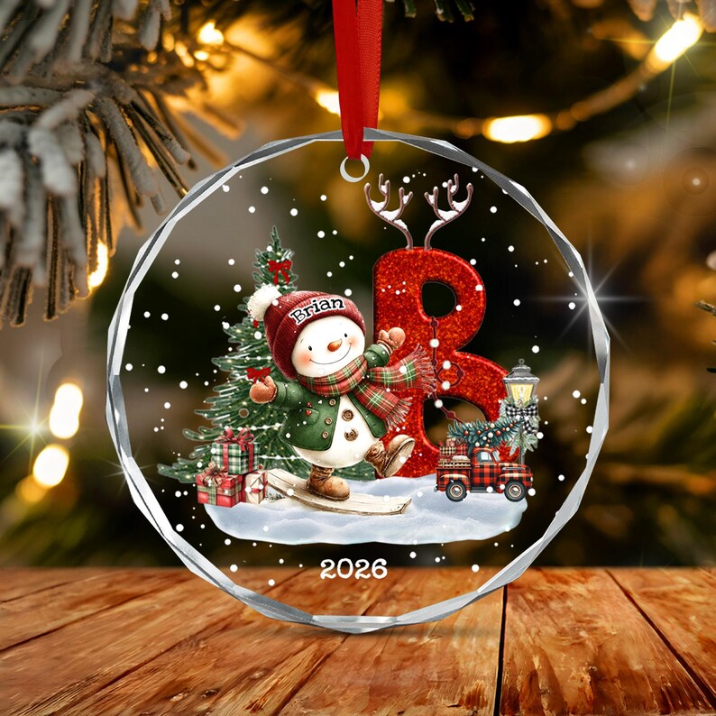 A circular clear acrylic Christmas ornament featuring a snowman wearing a red hat with the name Brian, a green coat, and a plaid scarf. A large red glittery letter B, a pine tree, gifts, a red truck, and the year 2026 are printed.