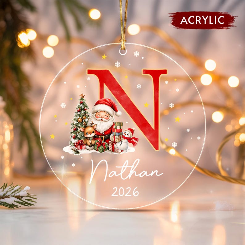 A round clear acrylic Christmas ornament featuring a large red letter N, a graphic of Santa Claus with a reindeer and snowman, the name Nathan, and the year 2026.