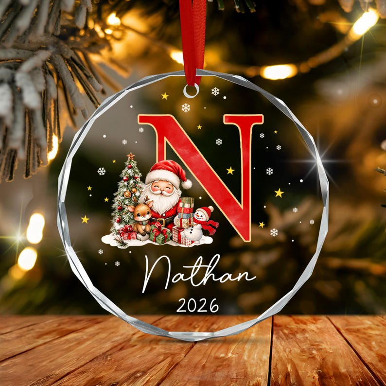A round, faceted glass Christmas ornament featuring a large red letter N, a cartoon Santa Claus, a reindeer, a snowman, and gift boxes.