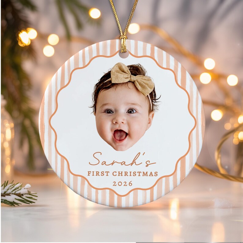 A round ceramic ornament featuring a photo of a baby wearing a tan bow headband. The background has vertical tan stripes and a scalloped border. Text reads: Sarah's FIRST CHRISTMAS 2026.