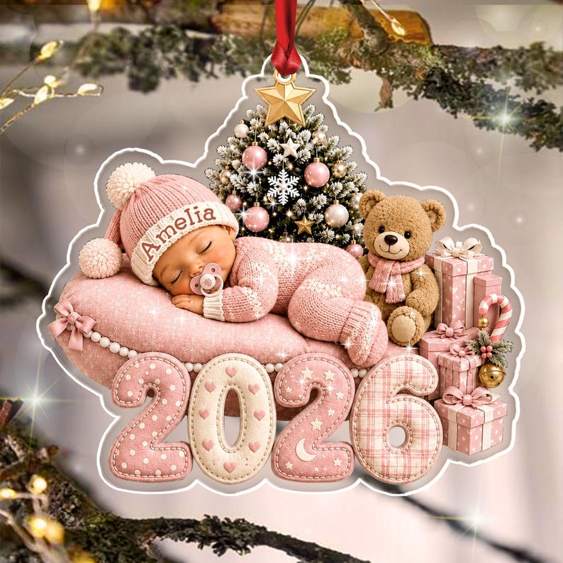 A decorative Christmas ornament featuring a sleeping baby in a pink knit outfit and hat with the name Amelia, a teddy bear, a small Christmas tree, and a stack of gifts, all positioned above the year 2026 in large.