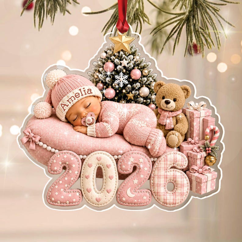 A decorative Christmas ornament featuring a sleeping baby wearing a pink knit hat with the name Amelia, resting on a pink pillow. Beside the baby is a teddy bear, a small Christmas tree, and gift boxes.