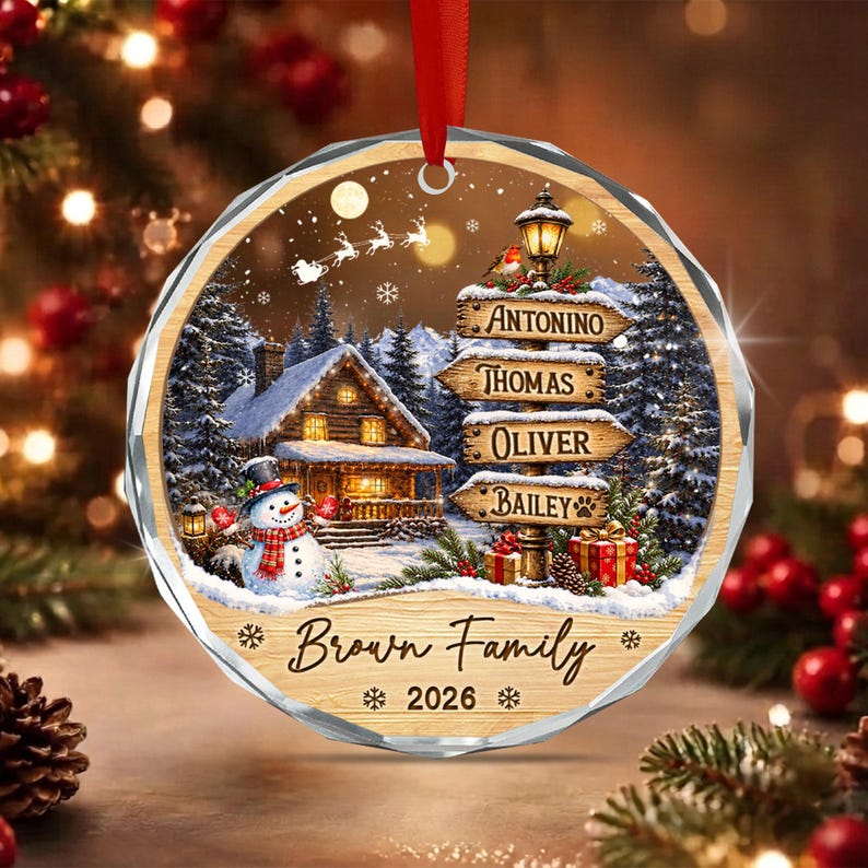 A round, faceted Christmas ornament featuring a snowy winter scene with a lit cabin, a snowman, and a signpost with the names Antonino, Thomas, Oliver, and Bailey. The text reads Brown Family and 2026, with a red ribbon hanger.