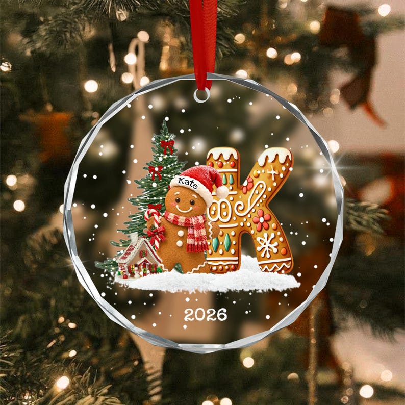 A round, clear glass Christmas ornament featuring a gingerbread person wearing a red hat with the name Kate, a red and white scarf, and a candy cane. Next to it is a large letter K, a small house, and the year 2026, all on snow.