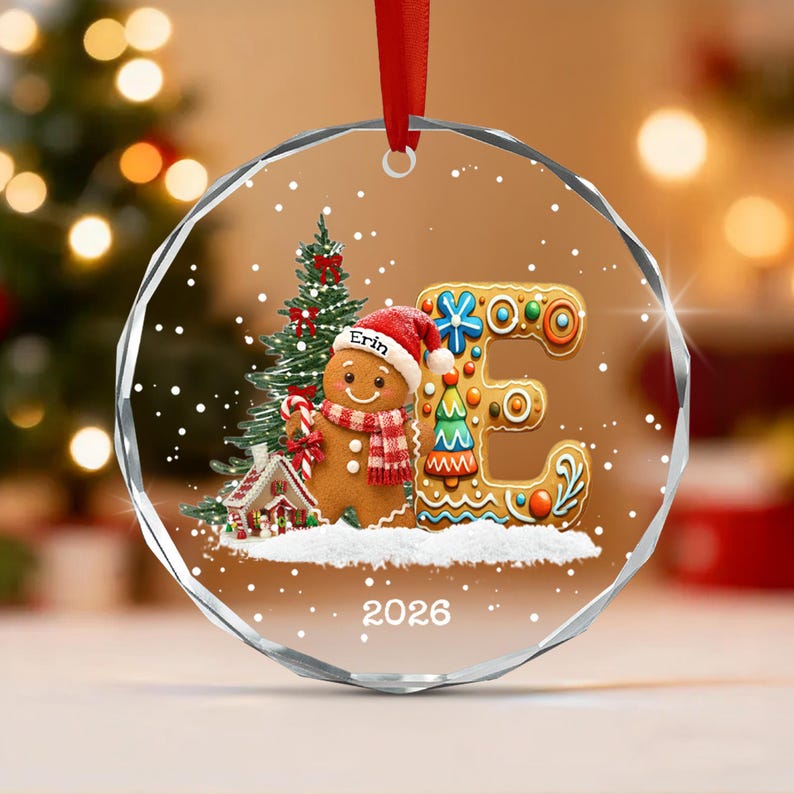 A circular clear glass Christmas ornament featuring a gingerbread person wearing a red hat with the name Erin, a red and white scarf, and holding a candy cane. Next to it is a large letter E, a pine tree, and the year 2026.