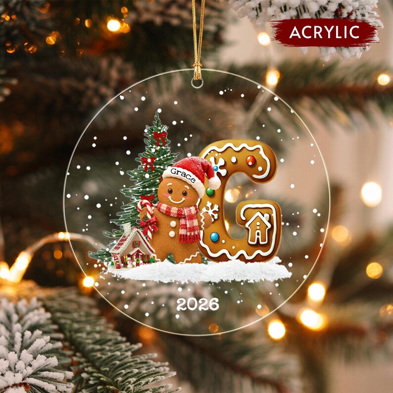 A round clear acrylic Christmas ornament featuring a gingerbread person wearing a red hat with the name Grace, a green Christmas tree, a small gingerbread house, a large letter G, and the year 2026.