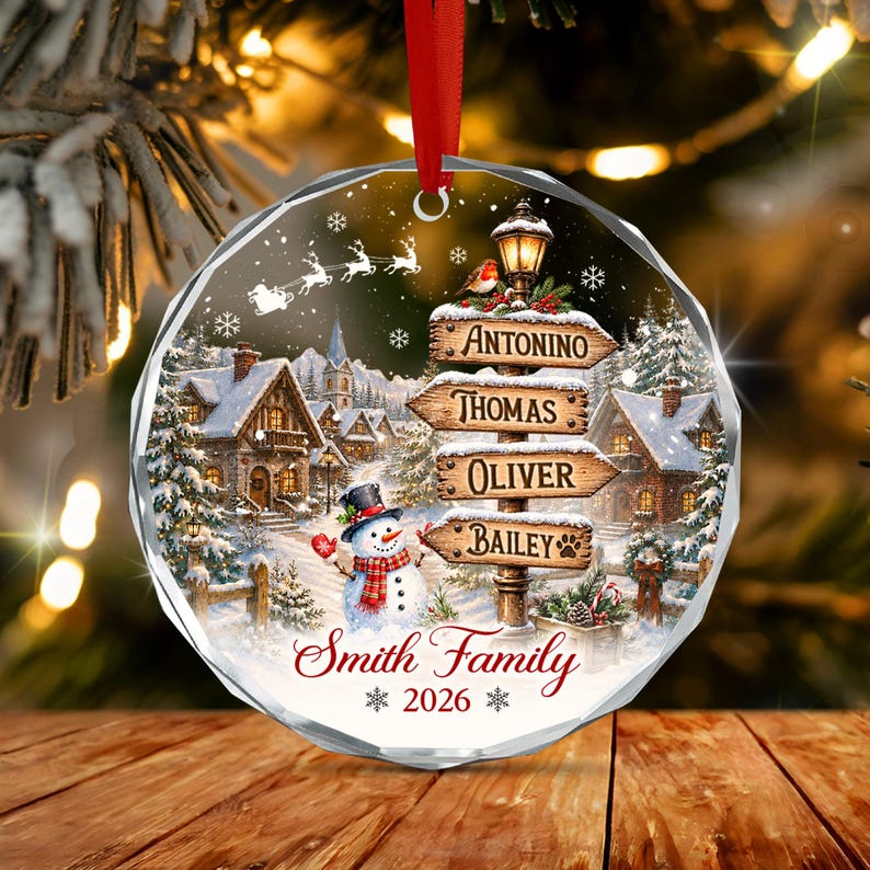A round, faceted glass Christmas ornament featuring a winter village scene with a snowman, a street lamp with four wooden signs labeled Antonino, Thomas, Oliver, and Bailey, and the text Smith Family 2026.