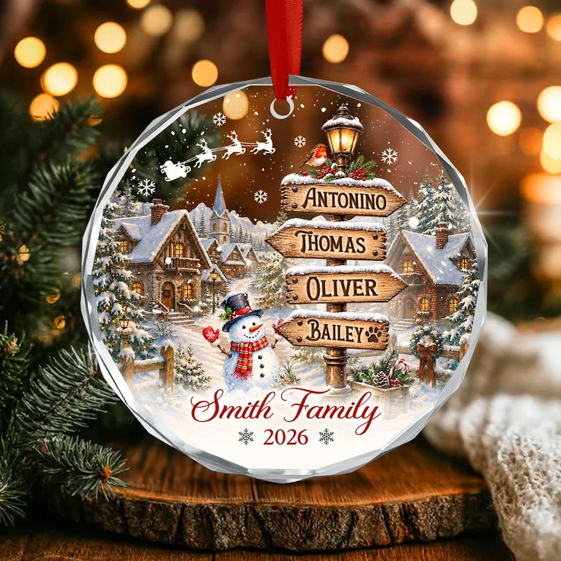 A round, faceted glass ornament featuring a winter village scene with a snowman, a signpost with the names Antonino, Thomas, Oliver, and Bailey, and the text Smith Family 2026. A red ribbon is attached to the top for hanging.