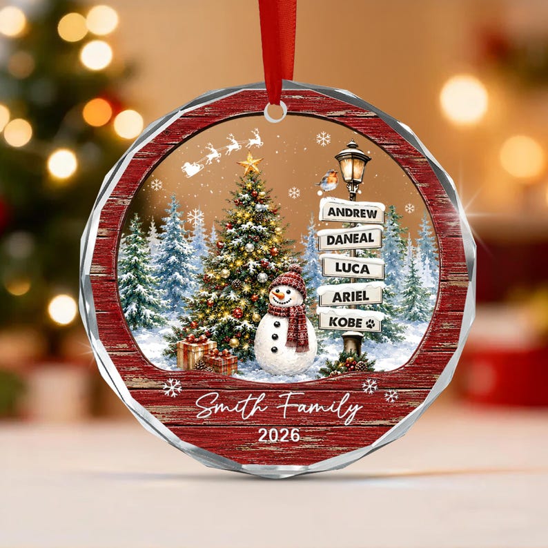 A round Christmas ornament with a red wood-grain border, featuring a snowy scene with a decorated tree, a snowman, and a street sign listing the names Andrew, Daneal, Luca, Ariel, and Kobe.
