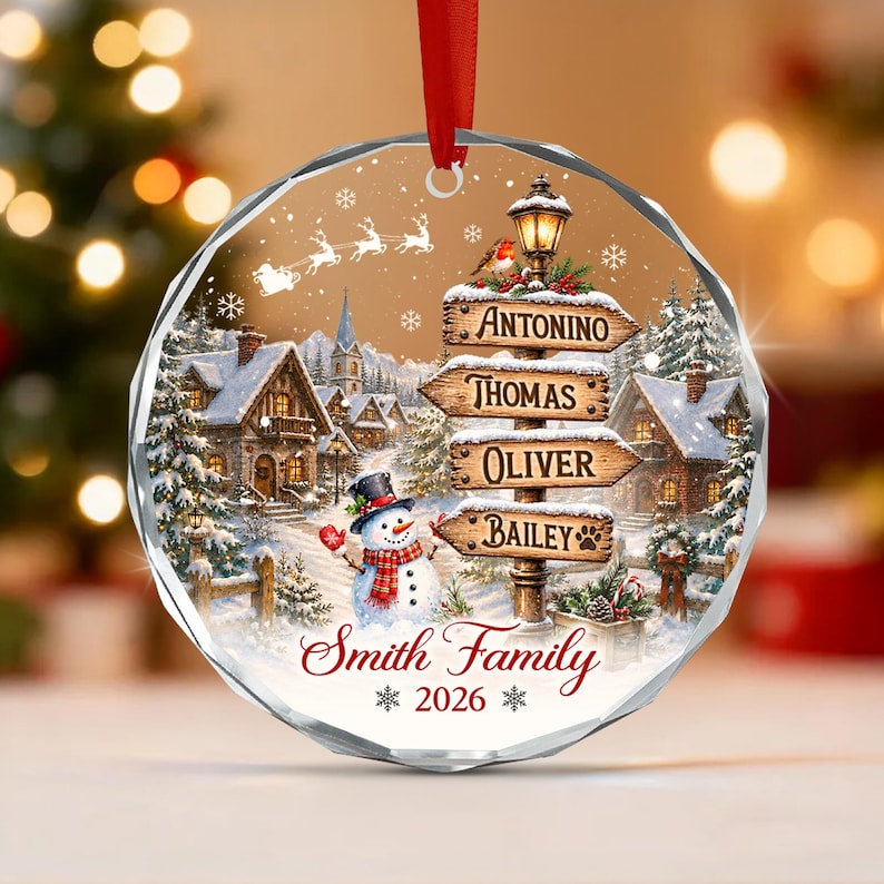 A circular glass Christmas ornament with a faceted edge and a red ribbon. The design features a snowy village, a snowman, a signpost with the names Antonino, Thomas, Oliver, and Bailey, the text Smith Family, and the year 2026.
