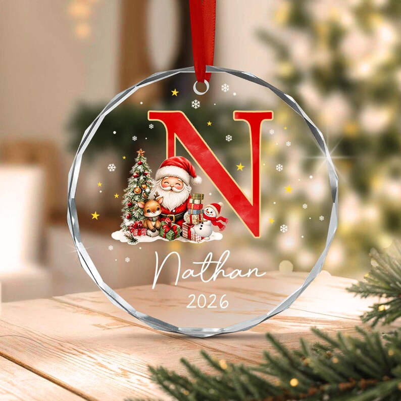 A round, clear, faceted glass Christmas ornament hanging from a red ribbon. It features a large red letter N, a graphic of Santa Claus with a reindeer and snowman, the name Nathan, and the year 2026 printed in white script text.