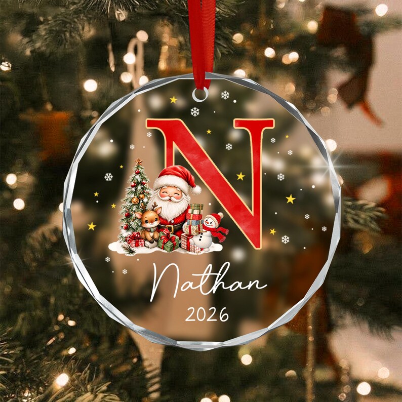 A round, clear acrylic Christmas ornament featuring a large red letter N, a graphic of Santa Claus with a reindeer and snowman, and the text Nathan 2026.