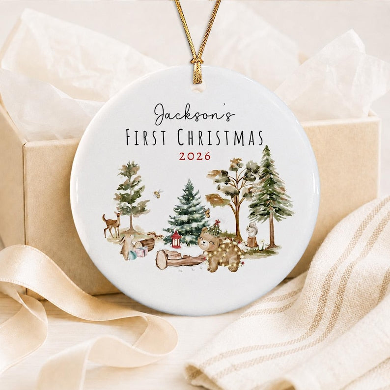 A round white ceramic ornament featuring a gold hanging string. The text reads Jackson's FIRST CHRISTMAS 2026. The design includes watercolor illustrations of a deer, a bear, a squirrel, pine trees, and a small red lantern.