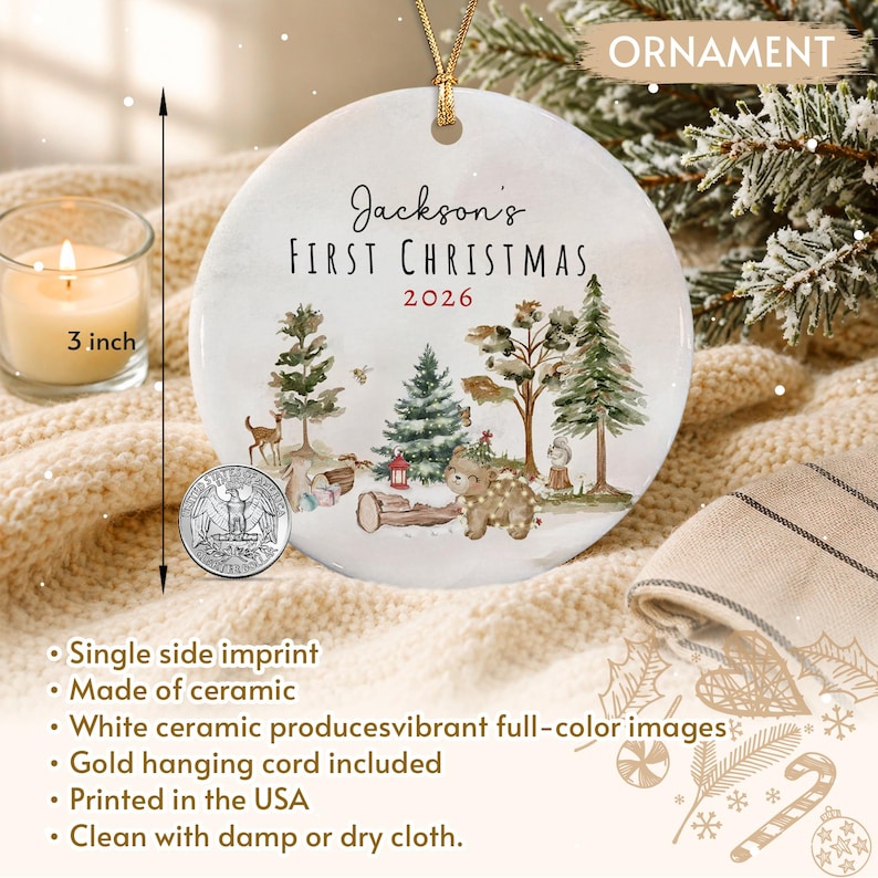 A round white ceramic ornament featuring the text Jackson's FIRST CHRISTMAS 2026 and a woodland scene with a bear, deer, and trees. A 3 inch measurement is indicated by a vertical arrow. A gold cord is attached for hanging.