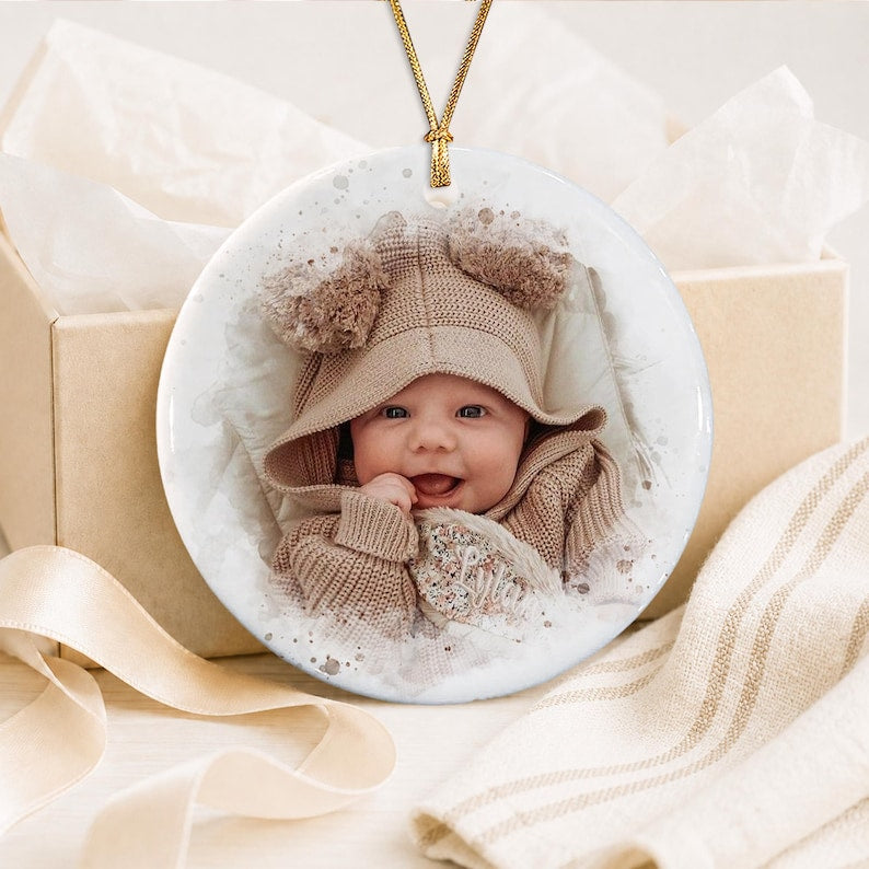 A round white ceramic ornament featuring a printed photograph of a smiling baby wearing a tan hooded knit sweater with bear ears. The ornament is suspended by a thin gold cord and displays the name Lyla in cursive script.