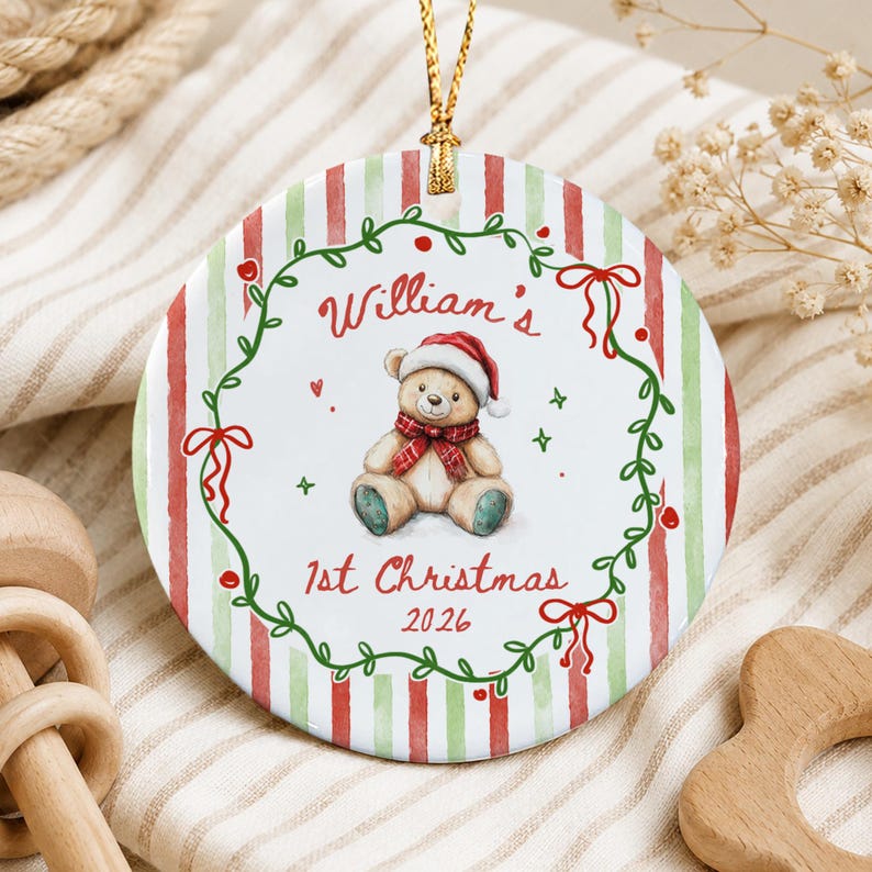 A round ceramic ornament featuring a red and green striped background. The center displays a teddy bear wearing a Santa hat and a red plaid scarf, surrounded by a green vine wreath with red bows.