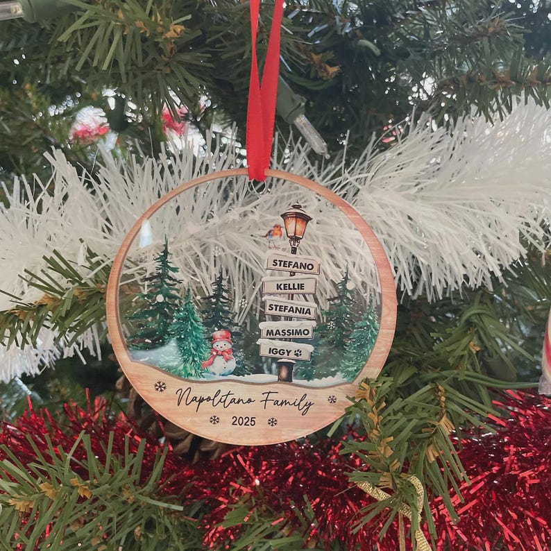 A round, flat, wooden Christmas ornament hanging by a red ribbon. The design features a snowy forest, a snowman, a street lamp with signs reading STEFANO, KELLIE, STEFANIA, MASSIMO, IGGY, and the text Napolitano Family 2025.