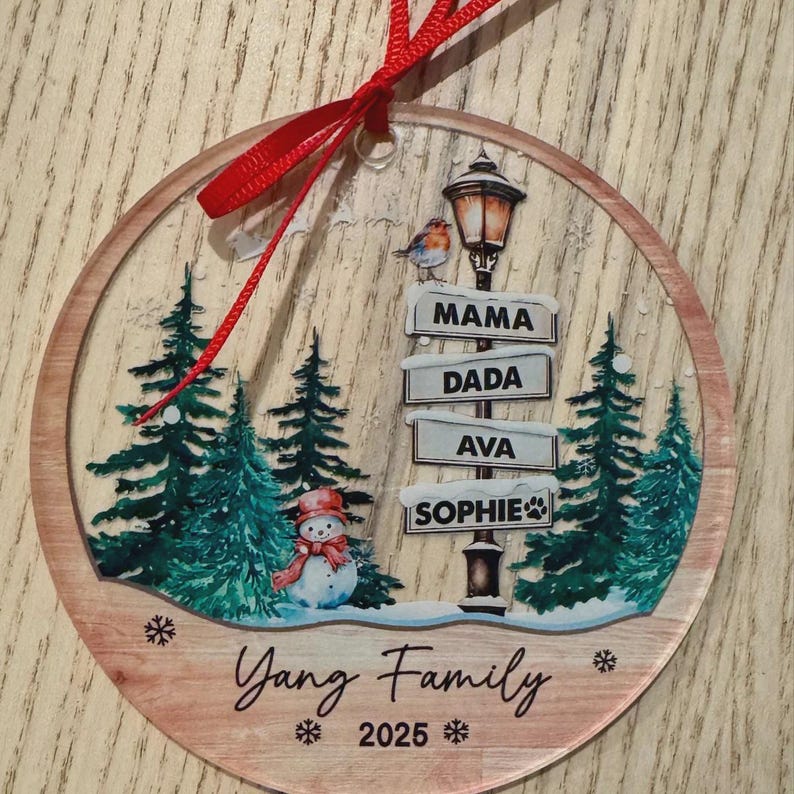 A round, clear acrylic ornament featuring a winter scene with green pine trees, a snowman, and a street lamp. The lamp post has signs reading MAMA, DADA, AVA, and SOPHIE.