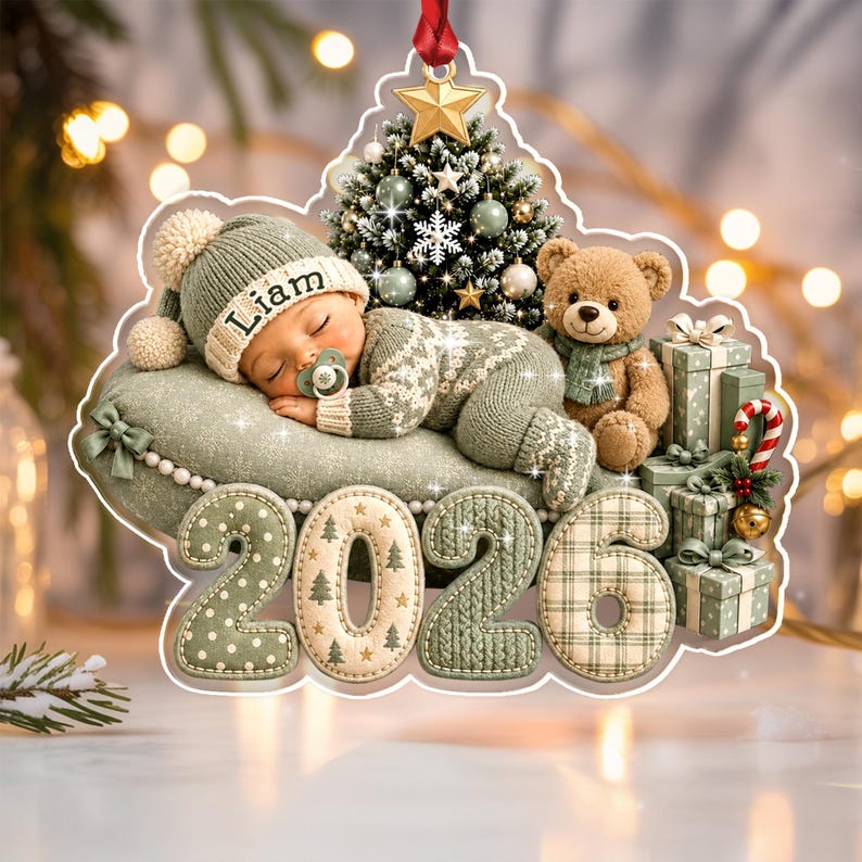 A Christmas ornament featuring a sleeping baby in a sage green knit outfit and hat with the name Liam, a teddy bear, a decorated tree, and gift boxes.