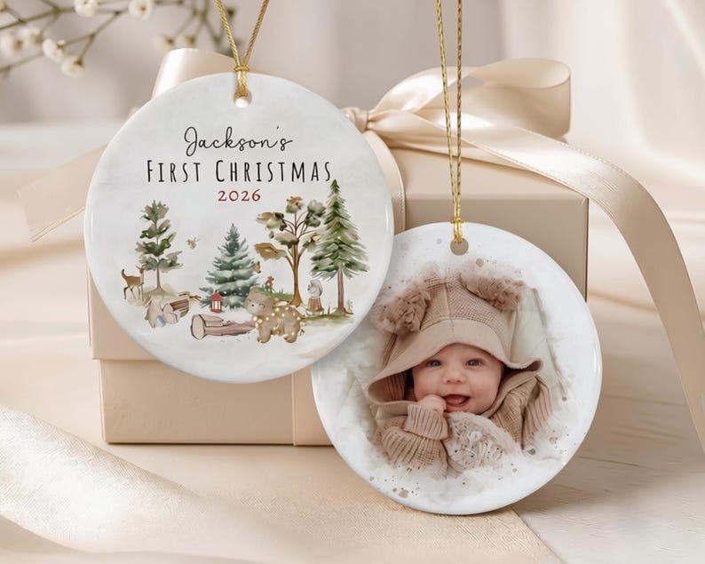 Two round white ceramic ornaments with gold hanging strings. One ornament features a watercolor forest scene with a deer, bear, and the text Jackson's First Christmas 2026.