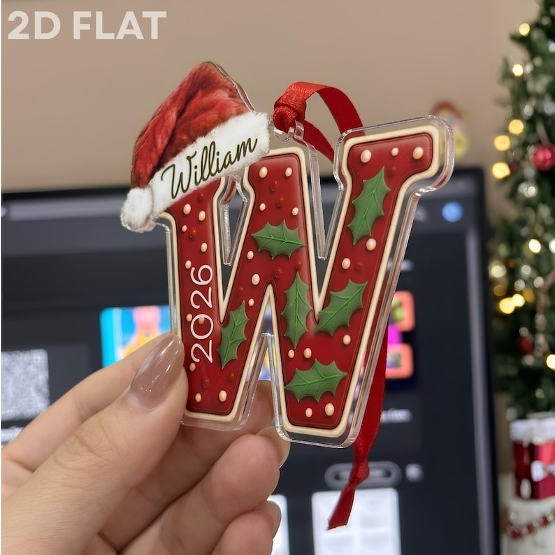 A clear acrylic Christmas ornament shaped like the letter W, featuring a red background with green holly leaves, white dots, and the year 2026. A red Santa hat sits atop the letter with the name William printed on the white trim.