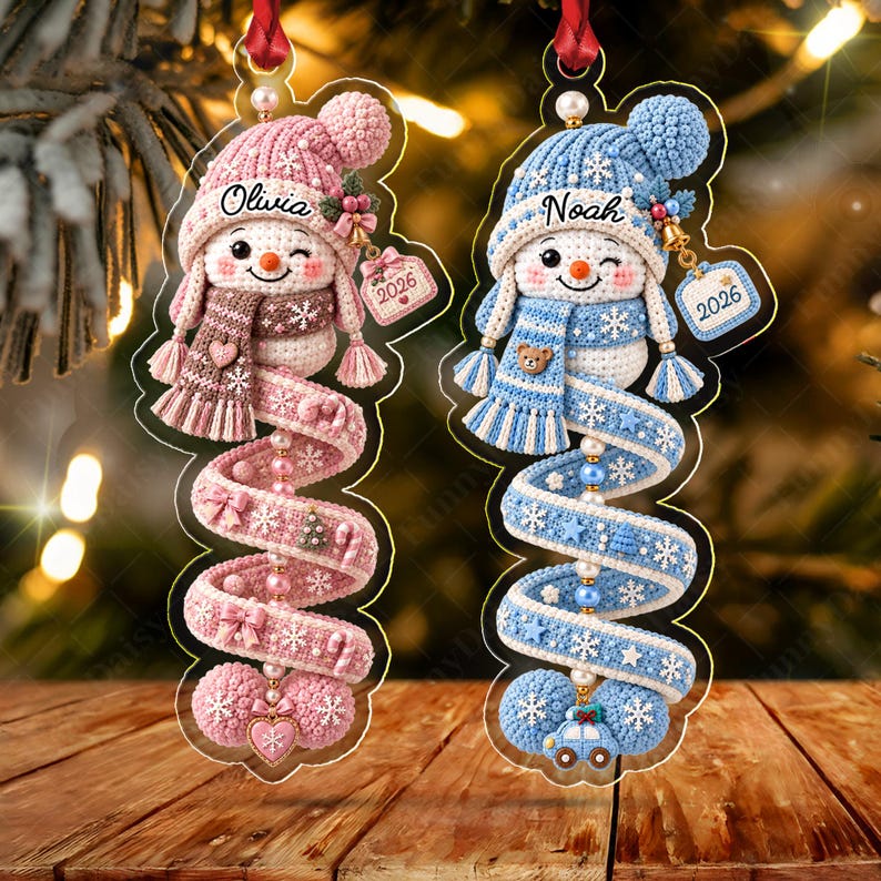 Two hanging ornaments featuring crocheted-style snowmen with pink and blue color schemes. The pink snowman is labeled Olivia and the blue one Noah, both with 2026 tags.