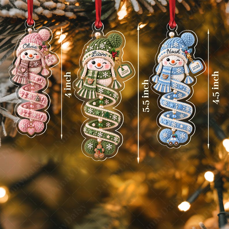Three hanging Christmas ornaments shaped like crocheted snowmen in pink, green, and blue. Each snowman wears a matching hat and scarf, with names Olivia, Liam, and Noah printed on the hats.