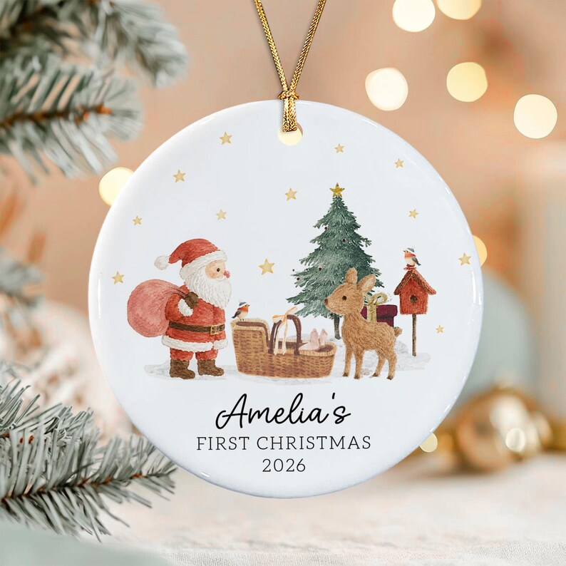 A round white ceramic ornament with a gold string featuring a watercolor illustration of Santa Claus, a reindeer, a Christmas tree, and a birdhouse. The text reads Amelia's FIRST CHRISTMAS 2026 in black lettering with gold stars.
