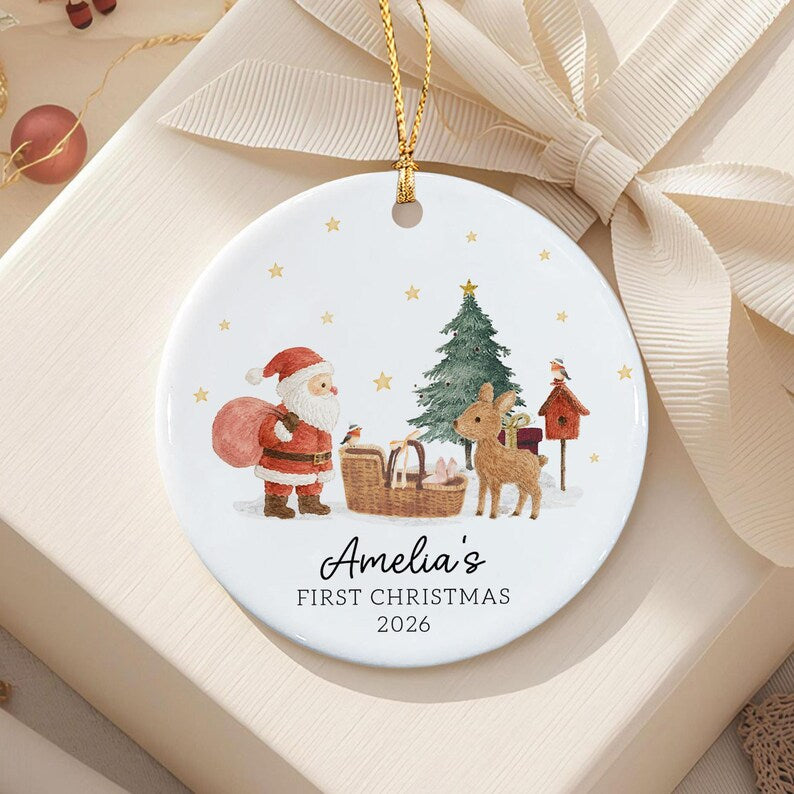 A round white ceramic Christmas ornament featuring a watercolor illustration of Santa Claus, a reindeer, a Christmas tree, and a birdhouse. The text reads Amelia's FIRST CHRISTMAS 2026.